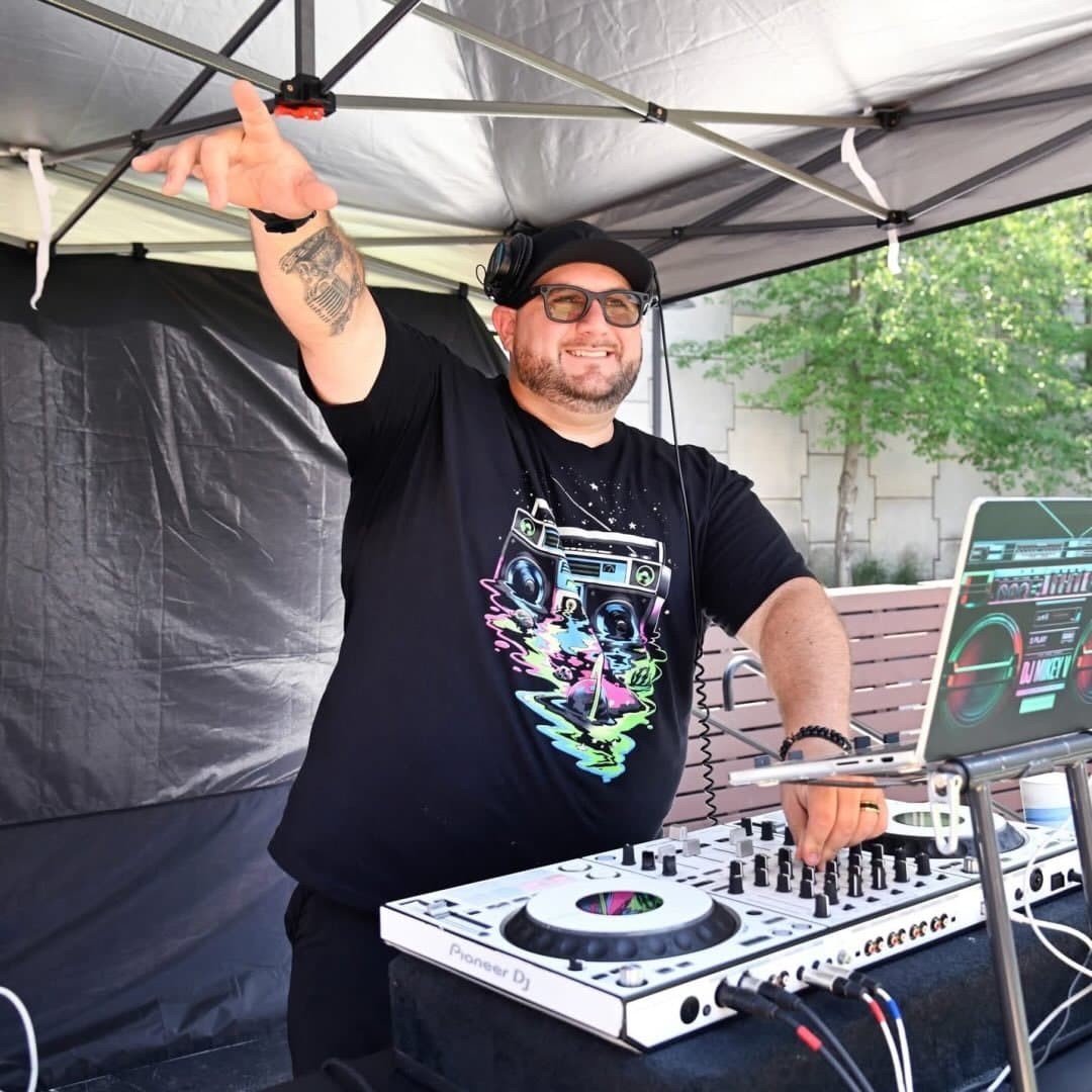 Smiling DJ in a black graphic tee points upward while mixing music on a white controller.