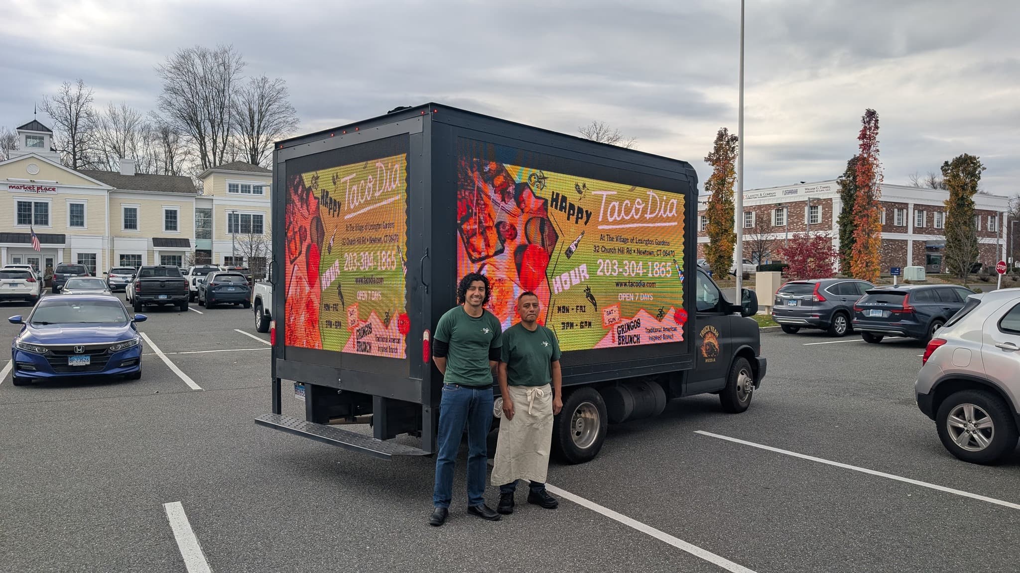 Two men in green shirts stand before a truck with a Taco Dia digital advertisement.