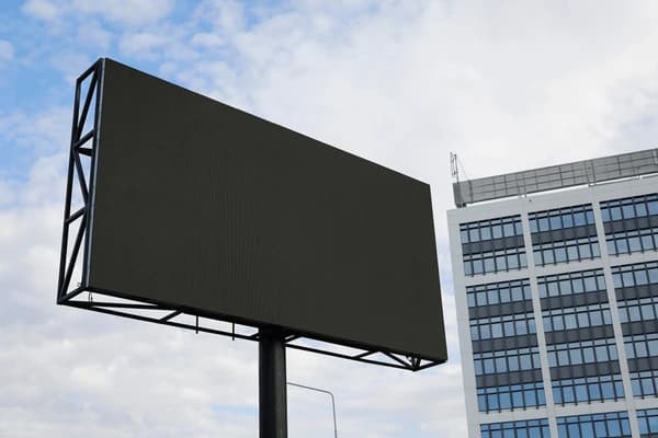 Blank black digital billboard on a metal frame against a cloudy sky and office building.